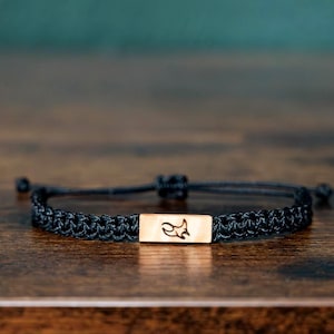 Pode incluir: Pulseira trançada preta com um detalhe retangular dourado com uma silhueta de raposa. A pulseira é ajustável e fica sobre uma superfície de madeira escura. O fundo é de cor turquesa desfocada.