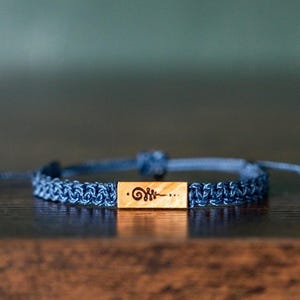 May include: A blue braided bracelet with a wooden rectangular centerpiece. The wooden piece has an etched design. The bracelet is adjustable with a sliding knot closure. The bracelet is resting on a dark wooden surface.