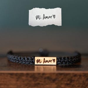 以下が含まれることがあります： 黒い編み込みコードのブレスレット。長方形のゴールドトーンの金属プレートには「Mi Amor」という文字とハートのシンボルが刻印されています。ブレスレットの上には、同じ文字とハートが黒いインクで書かれた白い紙が置かれています。