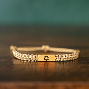 Op de afbeelding: Een beige macrame armband met een rechthoekig houten middelpunt. Het middelpunt heeft een zwart Davidster-symbool. De armband ligt op een donkere houten ondergrond met een vage turquoise en bruine achtergrond.