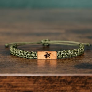 May include: An olive green braided cord bracelet featuring a rectangular wooden centre bead. The wooden bead is engraved with a small black line drawing of a sea turtle. The bracelet includes an adjustable sliding knot closure mechanism.