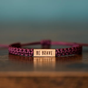 May include: A dark purple braided cord bracelet featuring a rectangular wooden centerpiece. The wooden piece is engraved with the text BE BRAVE in a simple sans-serif font. The bracelet has adjustable knotted ends and a woven texture.