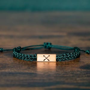 May include: A dark green woven cord bracelet featuring a rectangular wooden centerpiece. The wooden piece is engraved with a black graphic of two crossed arrows. The bracelet includes adjustable sliding knots on the cord for a secure fit.