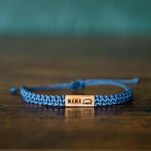 May include: Blue braided bracelet with a wooden rectangular accent. The wooden accent features the word "MAMA" and a bear silhouette. The bracelet is adjustable and sits on a wooden surface.