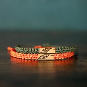 Könnte beinhalten: Zwei geflochtene Kordelarmbänder, eines in Olivgrün und eines in leuchtendem Orange, mit jeweils einer kleinen rechteckigen Holzperle, die mit einer schwarzen Strichzeichnung einer kletternden Person an einer Felswand graviert ist.