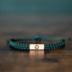 Puede incluir: Una pulsera de macramé verde oscuro con un acento rectangular de metal dorado. El acento metálico presenta un símbolo de triángulo dentro de un círculo. La pulsera está sobre una superficie de madera marrón oscuro.