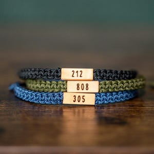 May include: Three braided bracelets in black, olive green, and blue, each with a small wooden tag. The tags display the numbers 212, 808, and 305. The bracelets are stacked on a wooden surface.