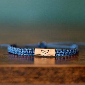Könnte beinhalten: Blau geflochtenes Armband mit einem rechteckigen Mittelstück aus Holz. Das Mittelstück zeigt ein Walflossendesign. Das Armband liegt auf einer Holzoberfläche mit verschwommenem blauem Hintergrund.