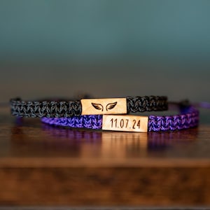 Op de afbeelding: Twee macram&eacute; armbanden, &eacute;&eacute;n zwart en &eacute;&eacute;n paars, met rechthoekige houten accenten. De zwarte armband heeft een vleugelontwerp en de paarse armband heeft de datum "11.07.24" gegraveerd.