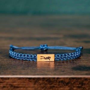 May include: A blue woven cord bracelet featuring a central rectangular light wood bead. The bead is engraved with a black silhouette of a scuba diver. The bracelet includes an adjustable sliding knot closure made from the same blue cord material.