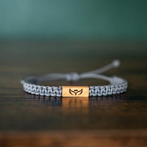 May include: A gray macrame cord bracelet featuring a central rectangular light brown wooden bead. The wooden bead is engraved with a black graphic design of a pair of wings with a small halo positioned directly above the center of the wings.