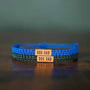 May include: Two braided cord bracelets, one in bright blue and one in dark green, each featuring a rectangular wooden bead engraved with the text DOG DAD. The bracelets are stacked horizontally, showcasing the woven texture and wood grain.