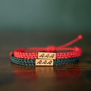 Può includere: Due braccialetti in corda intrecciata, uno rosso e uno verde scuro, ciascuno con una perla rettangolare in legno incisa con una grafica nera di tre piccoli uccelli seduti in fila, con estremità regolabili per una chiusura sicura al polso.