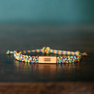 Puede incluir: Una pulsera con un cordón trenzado en un vibrante patrón de colores arcoíris. El centro de la pulsera está adornado con una pequeña cuenta de madera rectangular de color claro que muestra dos líneas horizontales paralelas grabadas en su frente.