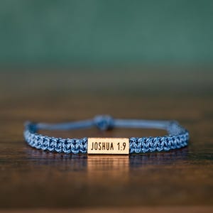 May include: Blue braided bracelet with a wooden rectangular accent. The wooden accent has the text "JOSHUA 1:9" engraved on it. The bracelet is on a dark brown wooden surface with a blurred green background.