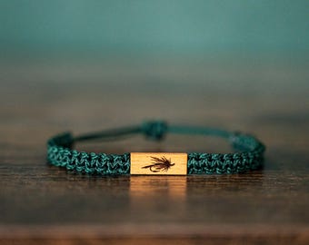 May include: A dark teal woven cord bracelet featuring a rectangular wooden centrepiece. The wooden piece is engraved with a detailed black illustration of a fishing fly. The bracelet has an adjustable sliding knot closure mechanism.