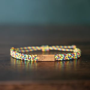 Puede incluir: Una pulsera que presenta un cordón trenzado con un patrón multicolor de hilos amarillos, azules, rojos y verdes. El centro de la pulsera incluye una cuenta de madera rectangular grabada con una serie de líneas horizontales y un patrón en chevron.