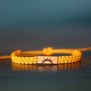 Puede incluir: Una pulsera de cordón amarillo con un dije rectangular de madera en el centro. El dije está grabado con un icono de sol negro. La pulsera está construida con un patrón de nudos trenzados e incluye cordones ajustables en ambos lados.