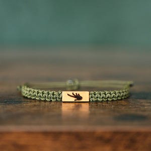 Peut inclure: Un bracelet en cordon vert olive doté d'une breloque rectangulaire en métal doré au centre. La breloque est gravée d'une silhouette sombre et détaillée d'une petite empreinte de main. Le cordon est tressé avec des extrémités réglables.