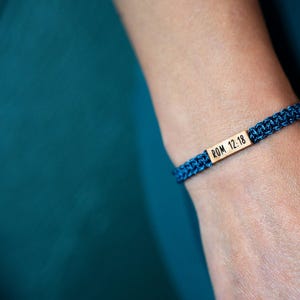 May include: A dark blue woven cord bracelet featuring a rectangular gold-toned metal bar centered on the band. The metal bar is engraved with the black text ROM 12:18. The bracelet has a braided texture and is secured around the wrist.