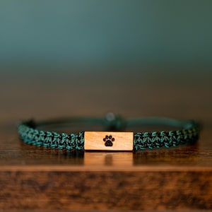 May include: A dark green woven cord bracelet featuring a rectangular light brown wooden centrepiece. The wooden piece is engraved with a small black paw print design centred on its surface. The bracelet has a simple, adjustable cord construction.