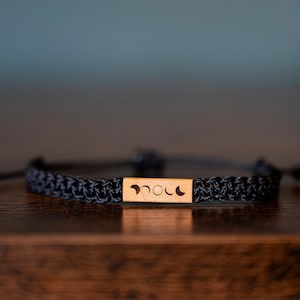 May include: A black braided cord bracelet featuring a rectangular light brown wooden centerpiece. The wooden piece is engraved with a series of five moon phase icons, showing the transition from a crescent moon to a full moon and back again.