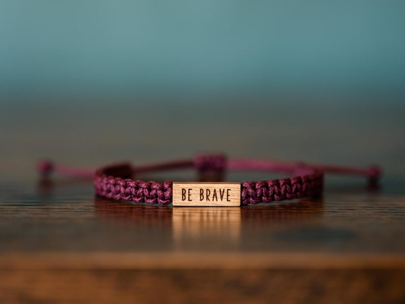 May include: A braided purple bracelet with a wooden rectangular accent piece. The wooden piece has the words "BE BRAVE" engraved on it. The bracelet is on a wooden surface with a blurred teal background.