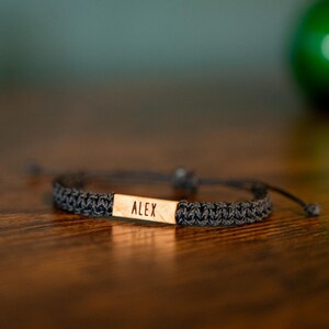 Op de afbeelding: Een donkergrijze gevlochten armband met een rechthoekig houten accent. Het woord "ALEX" is gegraveerd op het houten accent. De armband ligt op een donkerbruin houten oppervlak.