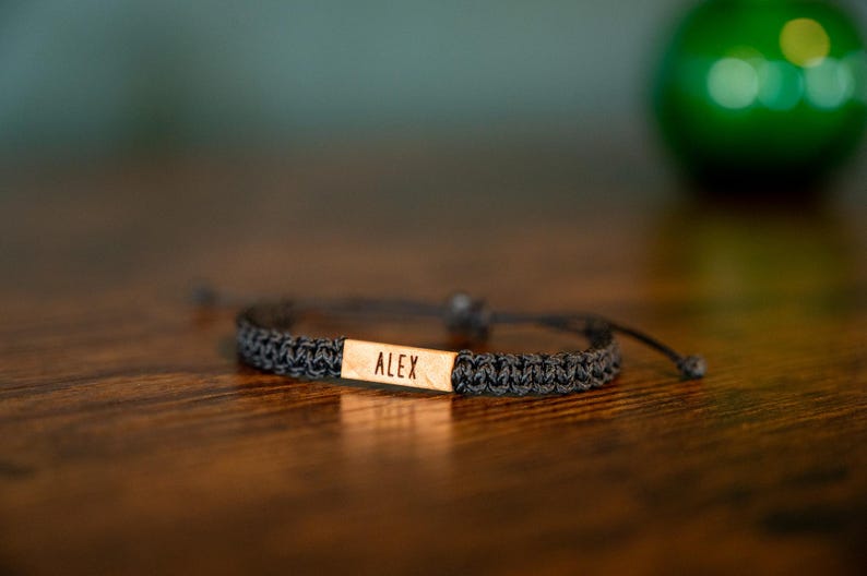 Op de afbeelding: Een donkergrijze gevlochten armband met een rechthoekig houten accent. Het woord "ALEX" is gegraveerd op het houten accent. De armband ligt op een donkerbruin houten oppervlak.