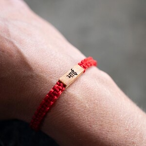 May include: A red braided cord bracelet featuring a rectangular light brown wooden centerpiece. The wooden piece is engraved with the black Devanagari script text "भाई", which translates to brother. The bracelet is secured with a woven knot design.