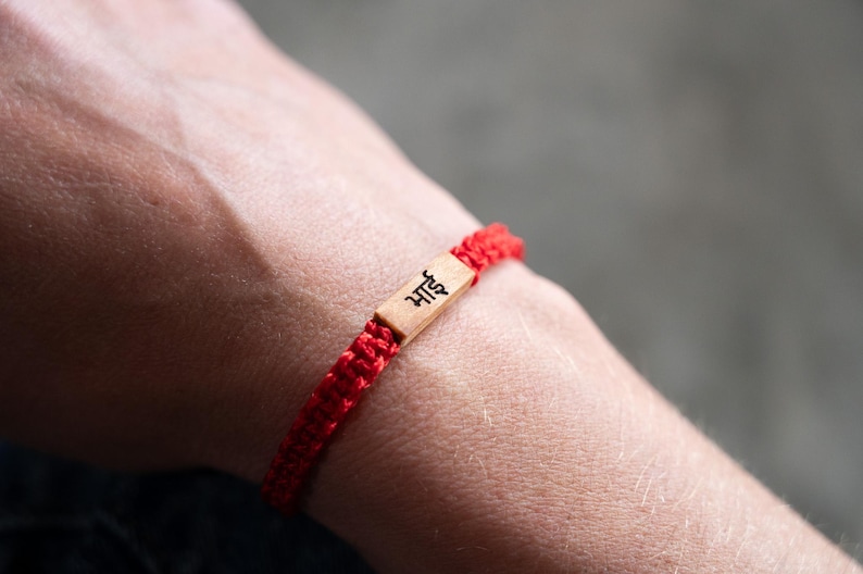 May include: A red braided cord bracelet featuring a rectangular light brown wooden centerpiece. The wooden piece is engraved with the black Devanagari script text "भाई", which translates to brother. The bracelet is secured with a woven knot design.