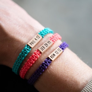 May include: Three braided cord bracelets in turquoise, coral, and purple. Each bracelet features a small, rectangular wooden bead engraved with black text reading PHIL 4:13, JER 29:11, and ROM 12:12, respectively, arranged in a stacked design.