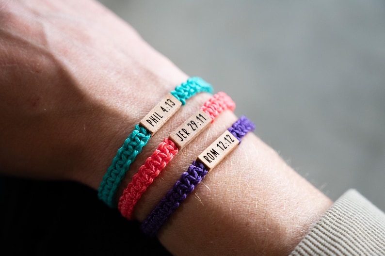 May include: Three braided cord bracelets in turquoise, coral, and purple. Each bracelet features a small, rectangular wooden bead engraved with black text reading PHIL 4:13, JER 29:11, and ROM 12:12, respectively, arranged in a stacked design.
