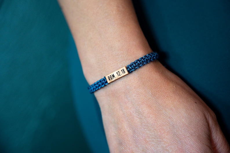 May include: A dark blue woven cord bracelet featuring a rectangular gold-toned metal bar centered on the band. The metal bar is engraved with the black text ROM 12:18. The bracelet has a braided texture and is secured around the wrist.