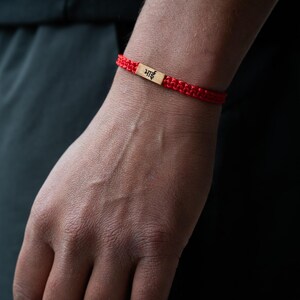 May include: A red woven cord bracelet featuring a rectangular gold-toned metal charm in the center. The charm is engraved with the Hindi word "भाई" in black lettering. The bracelet has a braided texture and is secured around the wrist.