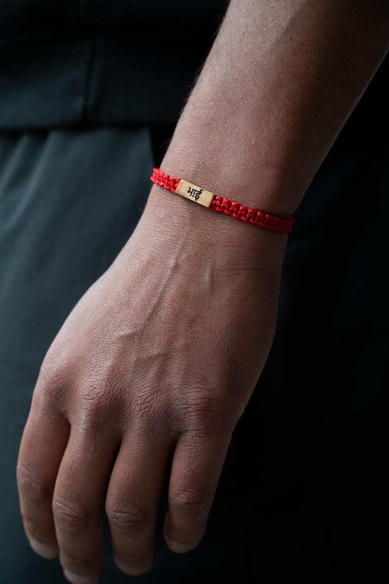 May include: A red woven cord bracelet featuring a rectangular gold-toned metal charm in the center. The charm is engraved with the Hindi word "भाई" in black lettering. The bracelet has a braided texture and is secured around the wrist.