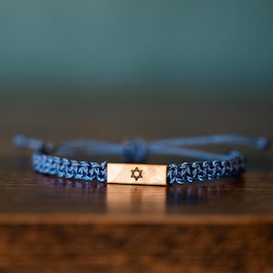 Op de afbeelding: Blauw geweven armband met een rechthoekig houten middelpunt. Het middelpunt heeft een Davidster-symbool. De armband is verstelbaar en ligt op een donkere houten ondergrond.