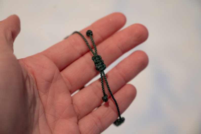 Op de afbeelding: Donkergroen koord met een glijknoop en twee decoratieve knopen aan de uiteinden. Het koord wordt in een hand gehouden tegen een witte achtergrond. Het koord lijkt gemaakt te zijn van een geweven materiaal.