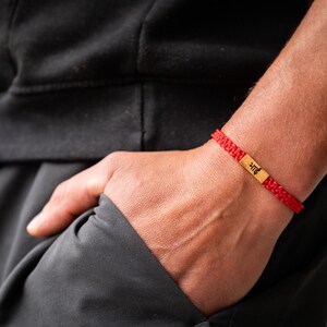 May include: A red woven cord bracelet featuring a rectangular gold-toned metal centerpiece. The metal plate is engraved with the black Hindi text "भाई". The bracelet has a braided texture and is secured around the wrist with a sliding knot closure.