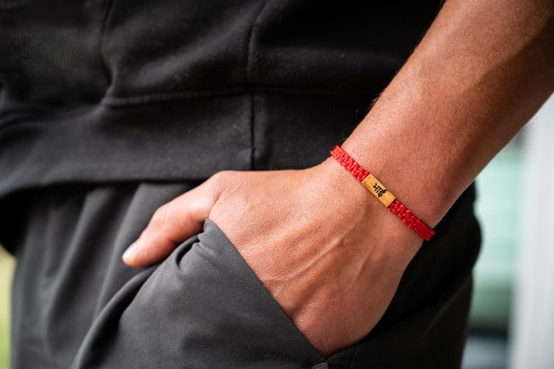 May include: A red woven cord bracelet featuring a rectangular gold-toned metal centerpiece. The metal plate is engraved with the black Hindi text "भाई". The bracelet has a braided texture and is secured around the wrist with a sliding knot closure.