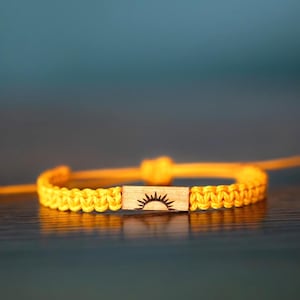 May include: A bright yellow woven cord bracelet featuring a rectangular wooden centerpiece. The wooden piece is engraved with a black graphic of a rising sun. The bracelet has adjustable pull strings on both sides to secure the fit around the wrist.