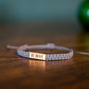 May include: A gray macrame bracelet with a gold rectangular charm that reads "BE BRAVE". The bracelet is adjustable and sits on a wooden surface. A green, blurred object is in the background.