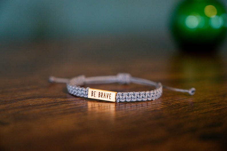May include: A gray macrame bracelet with a gold rectangular charm that reads "BE BRAVE". The bracelet is adjustable and sits on a wooden surface. A green, blurred object is in the background.