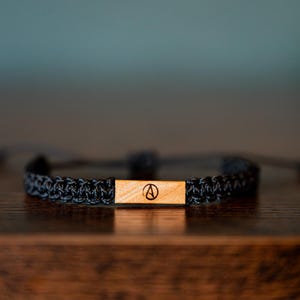 May include: A black woven cord bracelet featuring a rectangular light brown wooden centerpiece. The wooden plaque is engraved with a black circular symbol containing the letter A. The bracelet has adjustable cord ends for a secure fit.