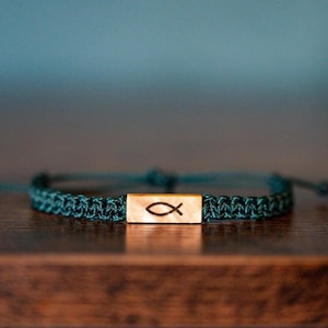 May include: A teal braided bracelet with a wooden centerpiece. The wooden piece features a carved fish symbol. The bracelet is adjustable and sits on a dark wooden surface.