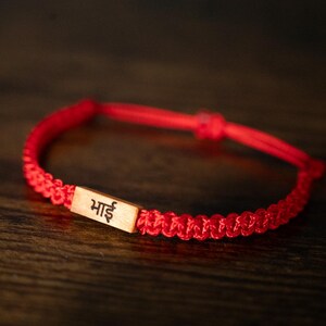 May include: A red braided cord bracelet featuring a rectangular light-colored wooden charm. The charm is engraved with the Devanagari script characters 'भाई' in black ink. The bracelet has an adjustable sliding knot closure mechanism.