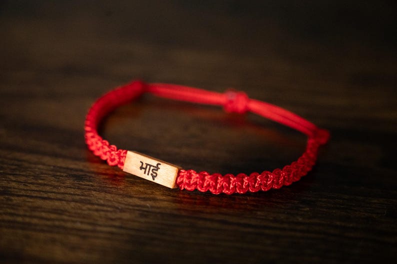 May include: A red braided cord bracelet featuring a rectangular light-colored wooden charm. The charm is engraved with the Devanagari script characters 'भाई' in black ink. The bracelet has an adjustable sliding knot closure mechanism.