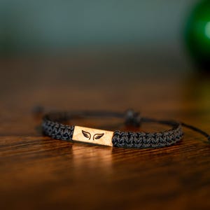 Op de afbeelding: Een zwart gevlochten armband met een rechthoekig houten middelpunt. Het middelpunt heeft een uitgesneden vleugelontwerp. De armband is verstelbaar en ligt op een houten oppervlak.