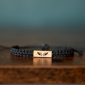 Op de afbeelding: Een zwart gevlochten armband met een rechthoekig houten middelpunt. Het middelpunt heeft een laserge&euml;tst ontwerp van twee vleugels. De armband is verstelbaar en ligt op een houten oppervlak.