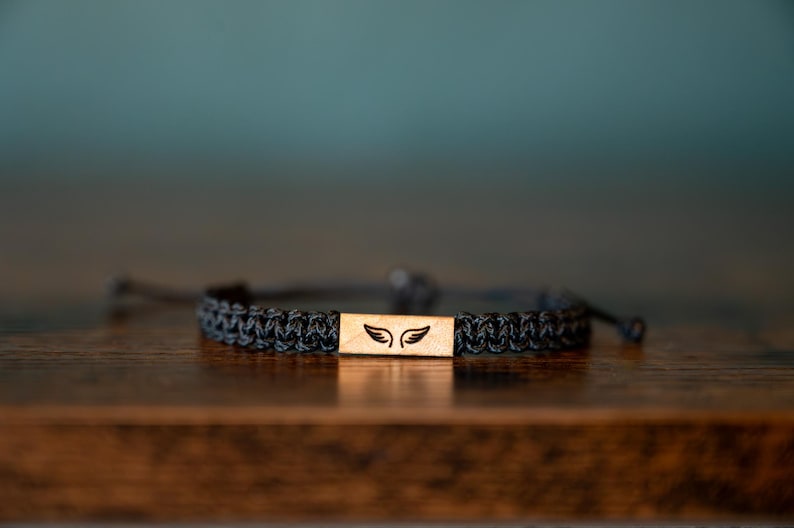 Op de afbeelding: Een zwart gevlochten armband met een rechthoekig houten middelpunt. Het middelpunt heeft een laserge&euml;tst ontwerp van twee vleugels. De armband is verstelbaar en ligt op een houten oppervlak.